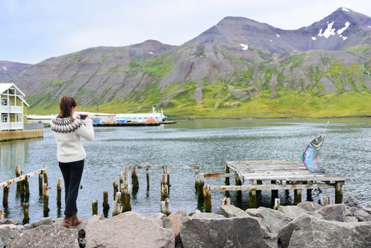 Tourist on Iceland taking photo of Siglufjordur Fjord wearing icelandic sweater. Woman visiting iceland sightseeing in Siglufjordur city taking picture using phone camera. North Iceland.