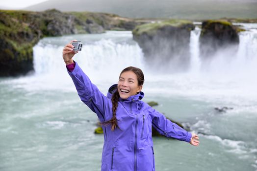 Iceland tourist taking selfie photo with smart phone by Godafoss waterfall. Happy young woman tourists enjoying icelandic nature landscape visiting famous tourist destination attraction, Iceland