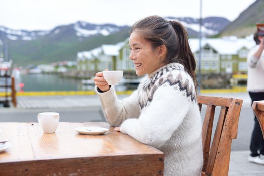 Woman drinking coffee on sidewalk cafe on Iceland. Girl tourist sipping enjoying coffee from cup wearing icelandic sweater. People visiting iceland sitting outdoors in Siglufjordur, North Iceland.