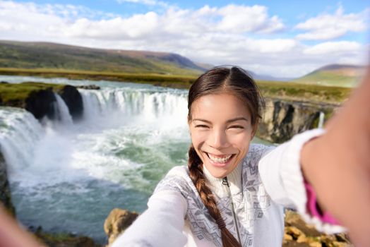 Tourist taking video selfie on travel by Godafoss waterfall on Iceland. Happy young woman tourists enjoying icelandic nature landscape visiting famous tourist destination attraction, Iceland.