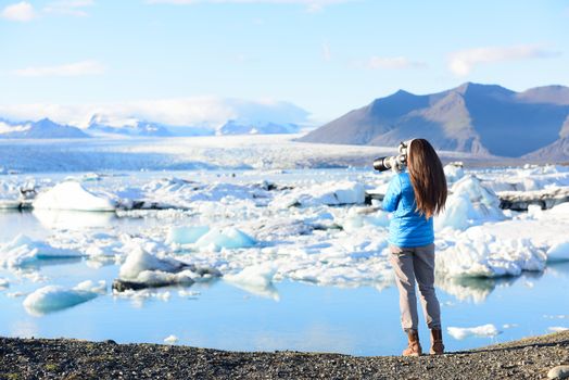 Photographer tourist woman taking photos with DSLR camera on travel on Iceland by Jokulsarlon glacial lagoon / glacier lake on Iceland. Happy tourist girl on travel in beautiful nature landscape.