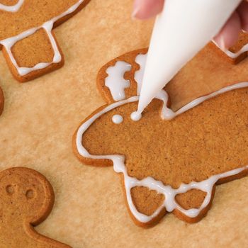 Young woman is decorating Christmas Gingerbread House cookies biscuit at home with frosting topping in icing bag, close up, lifestyle.