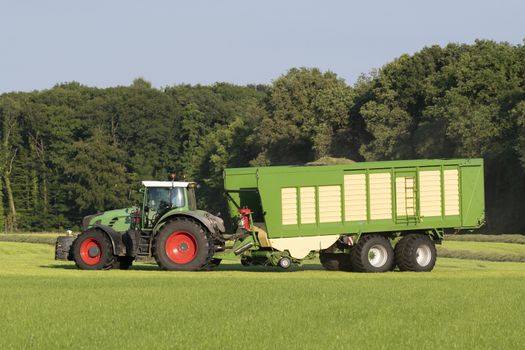 Green tractor picking up cut grass with a green loader wagon in the summer in the Netherlands