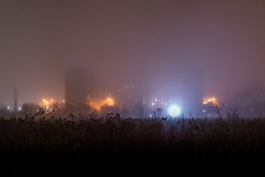 Dry dark grass field in front of foggy night depressive suburbs panel high-rise buildings ghetto with very shallow depth of field and selective focus.