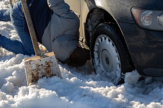 man working at car stuck in snow on knee with shovel at daylight offroad.