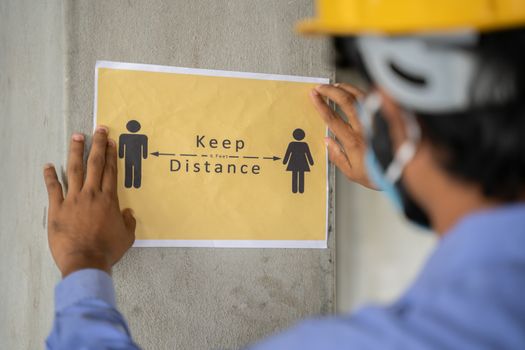 worker pasting Keep 6 feet distance on wall at work place or construction site due to coronavirus or covid-19 pandemic - concept of safety measures, back to work, new normal and protection