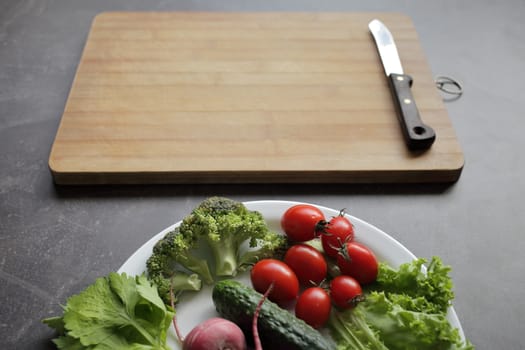 Fresh vegetables in a white plate on a gray table. Tomatoes, cucumber, lettuce, broccoli, radish, cutting board