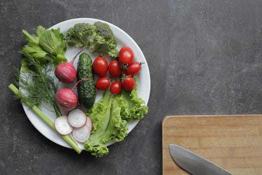 Fresh vegetables in a white plate on a gray table. Tomatoes, cucumber, lettuce, broccoli, radish, cutting board