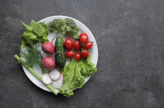 Fresh vegetables in a white plate on a gray table. Tomatoes, cucumber, lettuce, broccoli, radish
