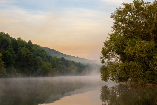 Misty calm small river on a summer morning. Fog floating over the cool water with reflection of the greenery and sky.