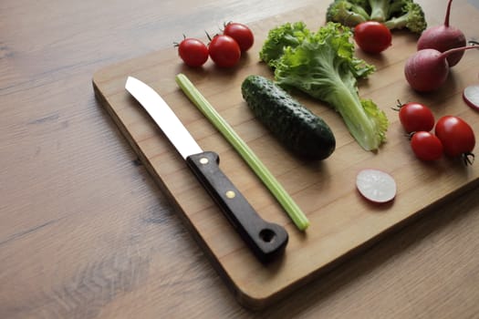 Fresh vegetables on a cutting board on a wooden table Tomatoes, cucumber, lettuce, broccoli, radish, knife