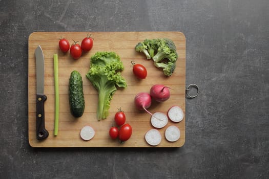 Fresh vegetables on a cutting board on a gray table Tomatoes, cucumber, lettuce, broccoli, radish, knife