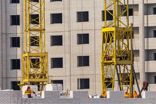 View of Construction Site With Unfinished Apartment Building, Cranes’ Bases and Construction Workers Dressed in Orange Outfit