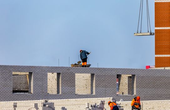 View of Construction Site With Workers Laying Bricks and Preparing Cement. Blue Sky and Unfinished Apartment Building Background.