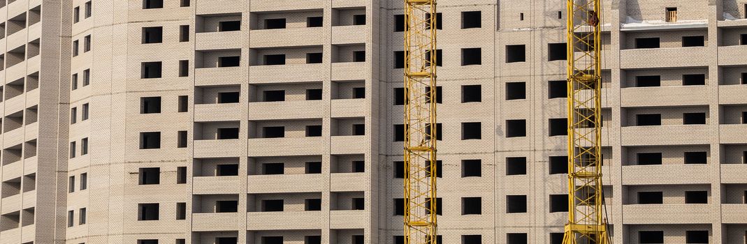 Construction site. Unfinished apartment building made of white bricks. Special yellow industrial construction cranes. Empty windows and balconies.