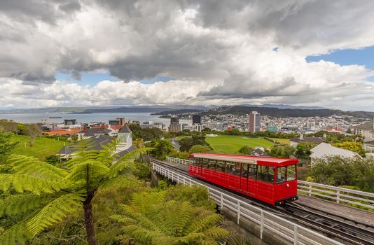 Red cable car moving up the mountain in the foreground and panorama of Wellington in the background. Beautiful blue sky covered with clouds.