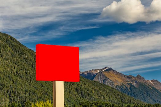 Blank red sign in the foreground and forest, mountains, blue sky with clouds in the background
