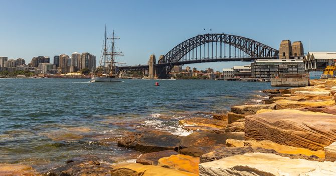 Panoramic view of Sydney harbor bridge and downtown buildings. Sailboat sailing in the harbor towards the bridge. Massive rocks in the foreground