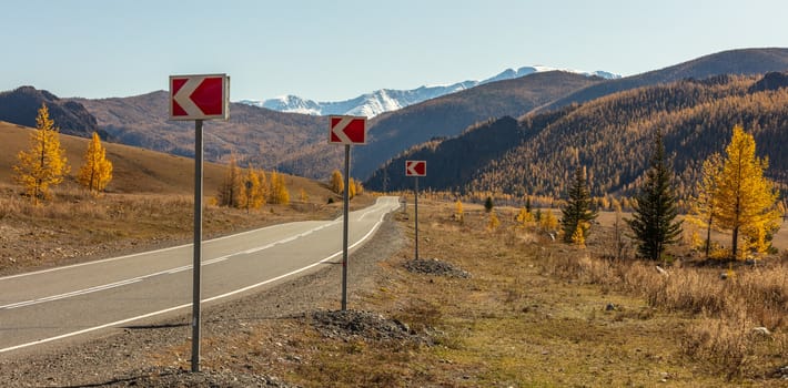 Panoramic view of a highway in Altai mountains. Red-and-white road signs on the curb with arrows pointing to the left. Snowy peaks in the background