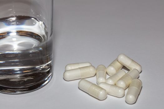 Close-up shot of pills and a half-full glass of water on white surface. Some of the pills and glass of water are is soft focus. Healthcare concept.