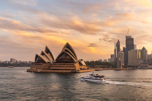 Sydney Harbor, Australia - November 1, 2018: Sydney Opera House at sunset. Beautiful orange-and-yellow colors. Down town and cloudy sky in the background. Tourist boat sailing the foreground.