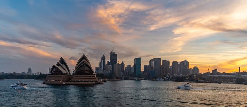 Sydney Harbor, Australia - November 1, 2018: Panorama. Sydney Opera House and downtown at sunset. Orange-and-yellow colors of the sky in the background. Tourist boats sailing in the foreground.