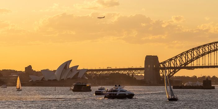 Sydney, Australia - November 30, 2018: Sydney Opera House and Harbor bridge at sunset. Beautiful orange-and-yellow colors of the cloudy sky in the background. Boats sailing.