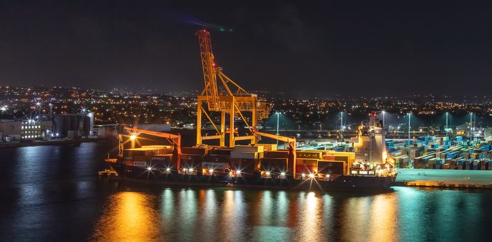 Bridgetown port, Barbados, West Indies - May 1, 2020: Night shot. Long exposure. Bridgetown port with loading crane and cargo ship being loaded with containers. Beautiful lights.