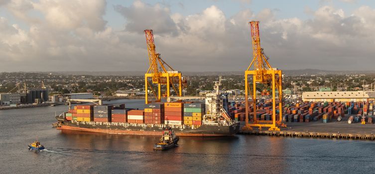 Bridgetown port, Barbados, West Indies - May 2, 2020: Bridgetown port with loading cranes and full cargo ship preparing to set sail. Tugboats ready to assist.