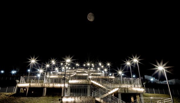 Night view of a staircase leading up to the top of the hill with the Moon above it and lights paving the way to the top. City of Barnaul, Siberia, Russia
