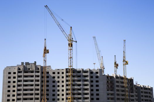 Distant view of a construction site with an unfinished apartment building and several cranes. Clear blue sky serves as a background.