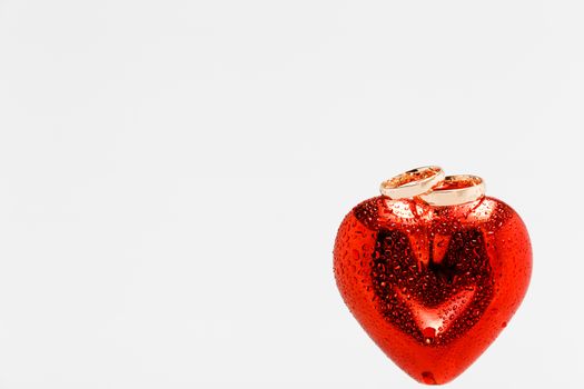 Two gold wedding rings on top of red heart with water drops all over them and the heart. Isolated on white background. Symbol of love and marriage.