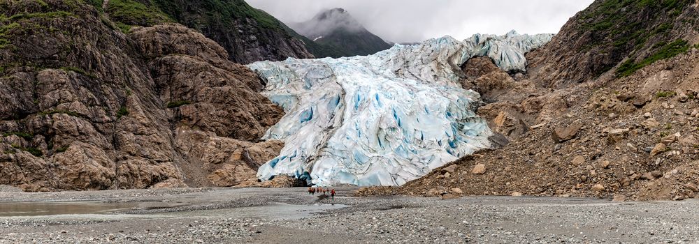 Panoramic view of Davidson Glacier in Alaska with tiny human figures approaching it in the distance