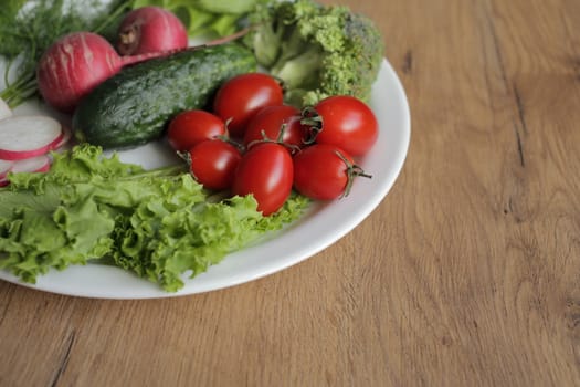 Fresh vegetables in a white plate on a wooden table. Tomatoes, cucumber, lettuce, broccoli, radish