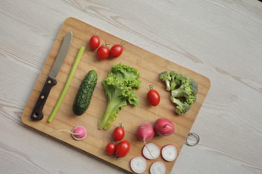 Fresh vegetables on a cutting board on a light wooden table Tomatoes, cucumber, lettuce, broccoli, radish, knife