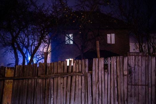 Dark foggy night view on wooden fence and spooky house surrounded by black dry apple trees with selective focus and shallow depth of field.