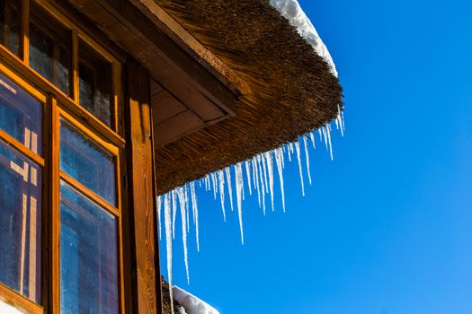 Icicles on straw roof corner of wooden rustic house with window