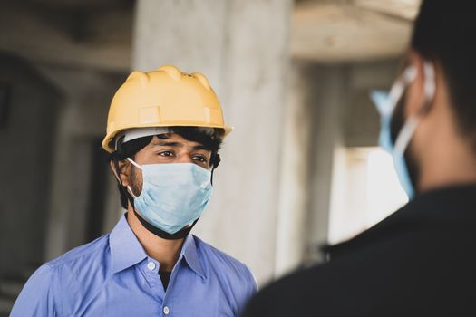 Two Construction workers or engineers at site talking by wearing medical face mask while maintaining social distance - concept of business, industry reopen or covid-19 safety measures at workplace