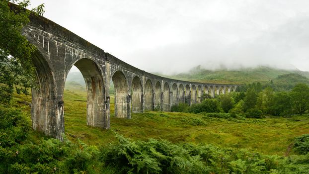 High resolution panorama of Glenfinnan railway viaduct (location from Harry Potter movie) shot from side, on overcast day with grey sky and lot of green grass and trees around. Inverness, Scotland