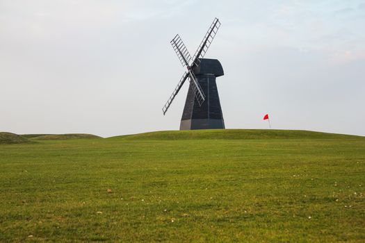 Windmill and red flag with green lawn and overcast sky in Rottingdean Windmill golf course. East Sussex, England.