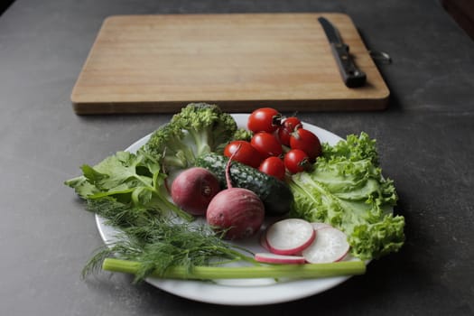 Fresh vegetables in a white plate on a gray table. Tomatoes, cucumber, lettuce, broccoli, radish, cutting board