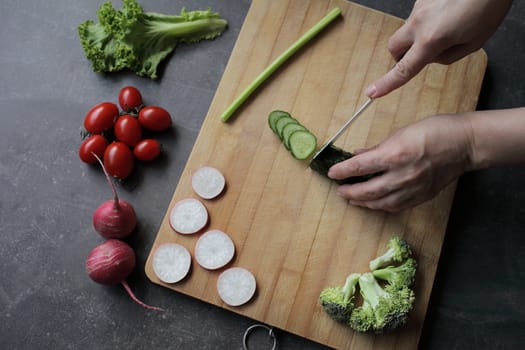 Hands cut cucumber on a cutting board on a gray table. Fresh vegetables, tomatoes, lettuce, broccoli, radishes lie nearby