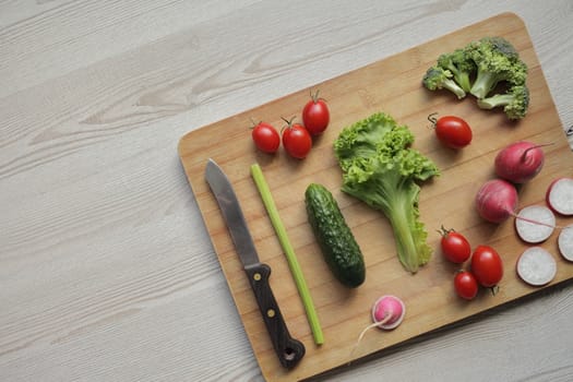 Fresh vegetables on a cutting board on a light wooden table Tomatoes, cucumber, lettuce, broccoli, radish, knife
