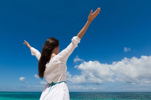 Woman in white dress posing in tropical sea beach with arms raised