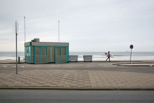 Lone surfer in neoprene swimsuit walking on pavement to empty beach on a cold winter overcast day. The Hague, Netherlands