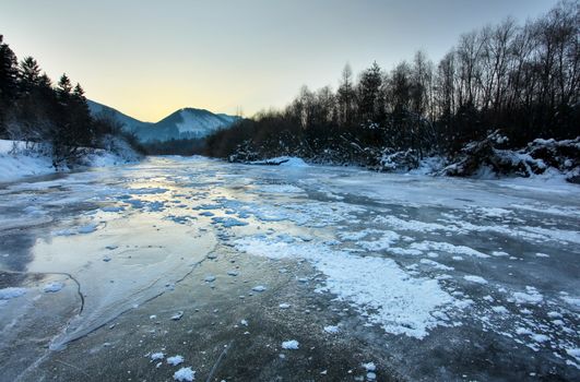 River Bela completely frozen during extreme cold, early morning sun reflected in ice layer covered with snow patches. Liptovsky Hradok, Slovakia