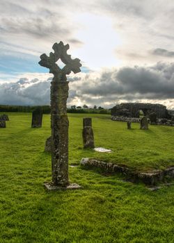 Old celtic stone cross on cemetery full of green grass with some ruins and sun creating backlight through clouds in background. Devenish Island, Enniskillen, Northern Ireland