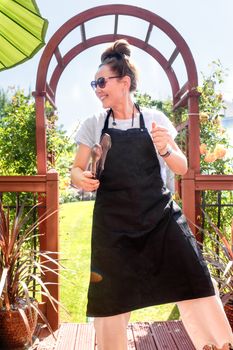 A young woman in a black apron making a barbecue and dancing on a garden