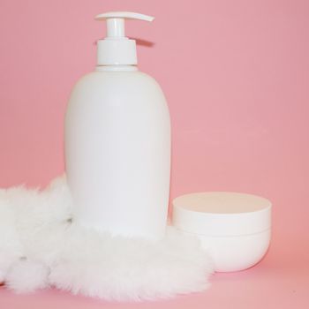 white cosmetic bottle, cream jar and white fur on pink background close-up