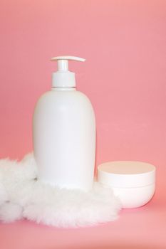 white cosmetic bottle, cream jar and white fur on pink background close-up
